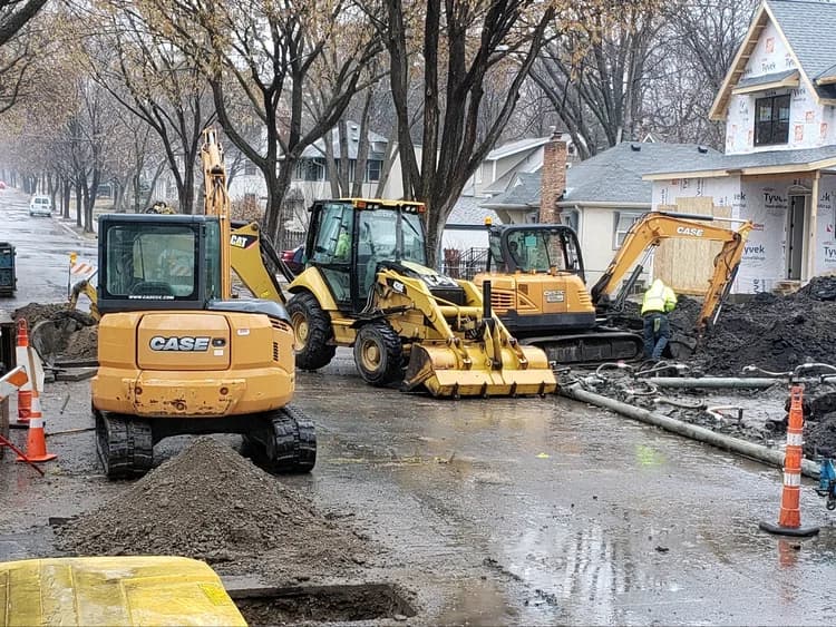 Yellow excavators, a backhoe, and worker on a muddy street construction site.