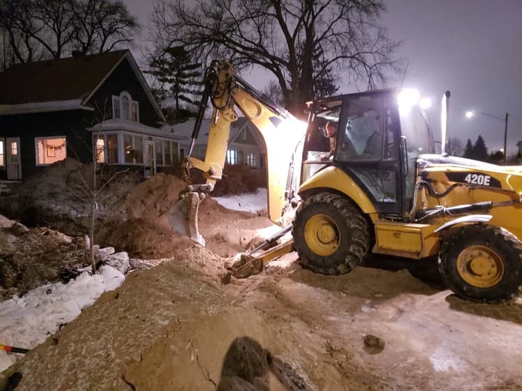 Yellow backhoe digging a trench at night, operator visible, on snowy ground near a house.