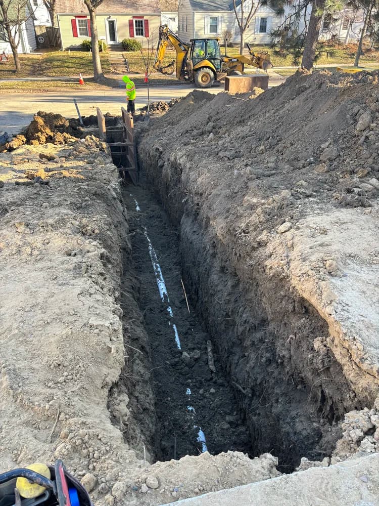 Long, deep trench with new pipe, worker, backhoe, and dirt piles in a neighborhood.